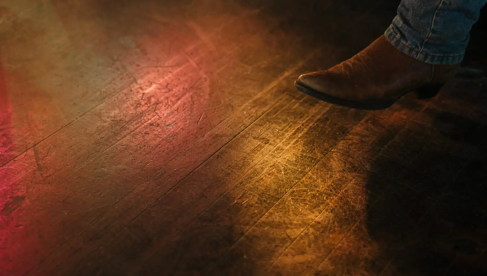 Un piso de madera para bailar lleno de marcas de botas, con la punta de una bota vaquera café en el borde y luz roja y ámbar del escenario