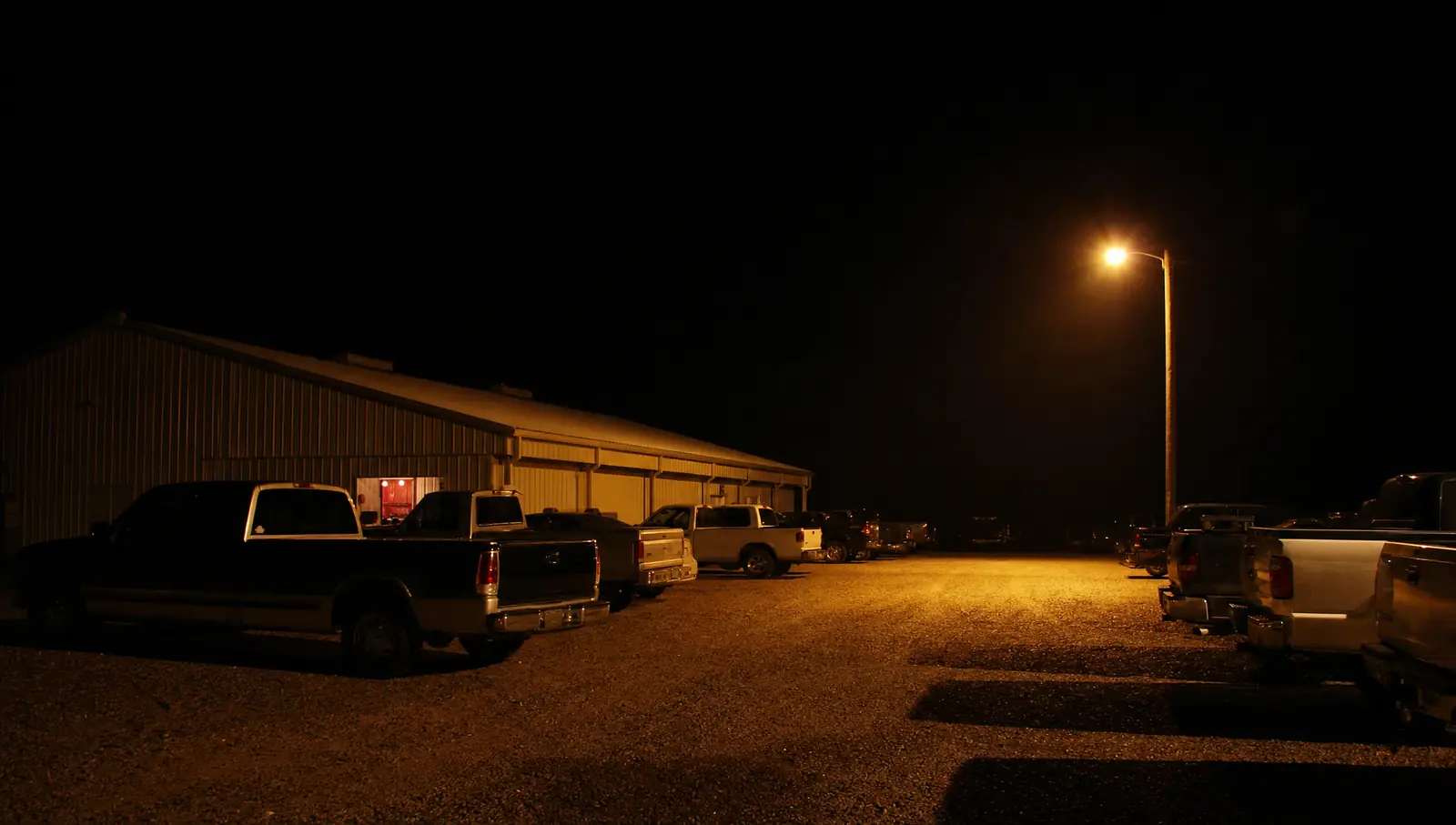 Un estacionamiento de grava de noche frente a un salón de baile con techo de lámina, filas de camionetas y un solo poste de luz anaranjada
