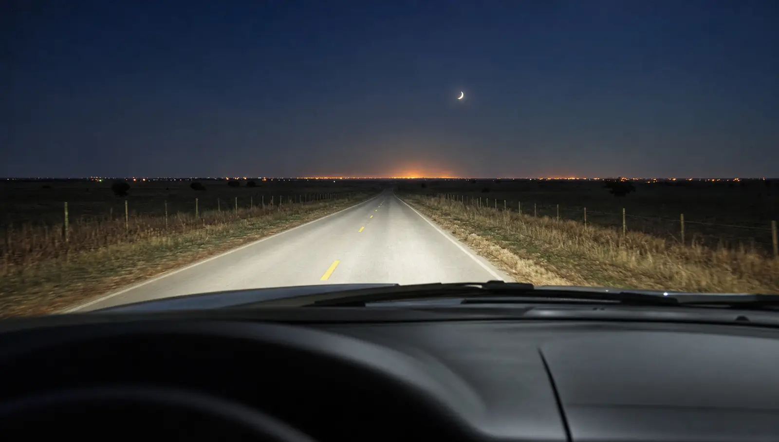 Una carretera de dos carriles de noche con la línea central amarilla, las luces traseras de una camioneta a lo lejos y el resplandor de una ciudad en el horizonte
