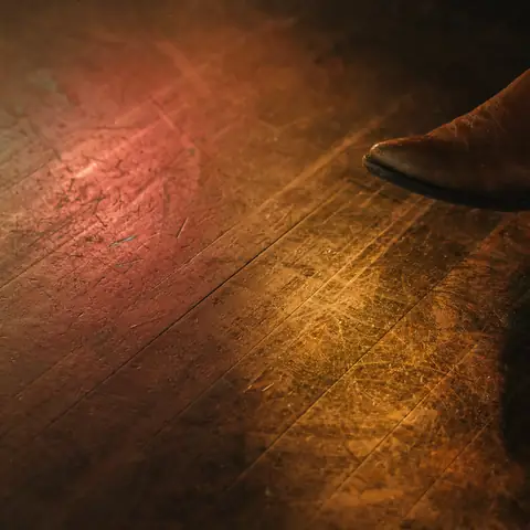 Un piso de madera para bailar lleno de marcas de botas, con la punta de una bota vaquera café en el borde y luz roja y ámbar del escenario