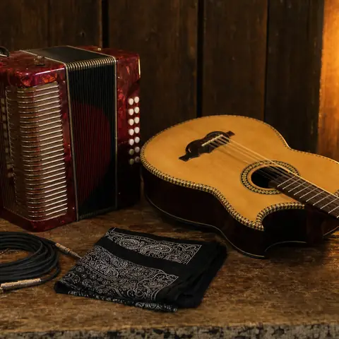 Un acordeón rojo de botones y una guitarra de cuerdas dobles sobre una mesa de madera detrás del escenario, junto a un cable y un paliacate, bajo la luz de un foco