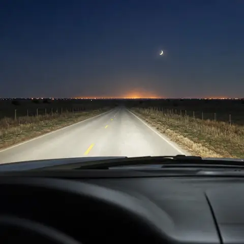 Una carretera de dos carriles de noche con la línea central amarilla, las luces traseras de una camioneta a lo lejos y el resplandor de una ciudad en el horizonte