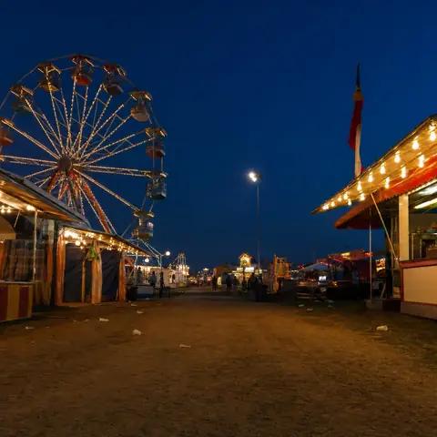 El paseo de una feria de pueblo al anochecer, con una rueda de la fortuna iluminada y puestos de comida con toldos encendidos