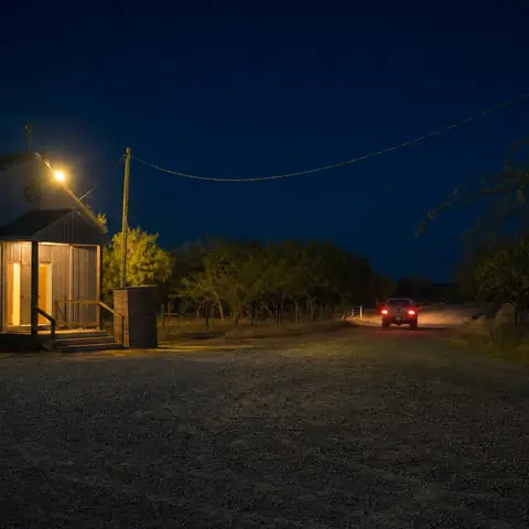 Una camioneta con las luces encendidas en una carretera vacía frente a un salón de baile rural, de noche, con un poste de luz anaranjada