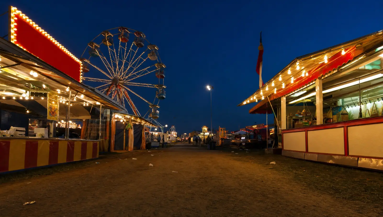 El paseo de una feria de pueblo al anochecer, con una rueda de la fortuna iluminada y puestos de comida con toldos encendidos