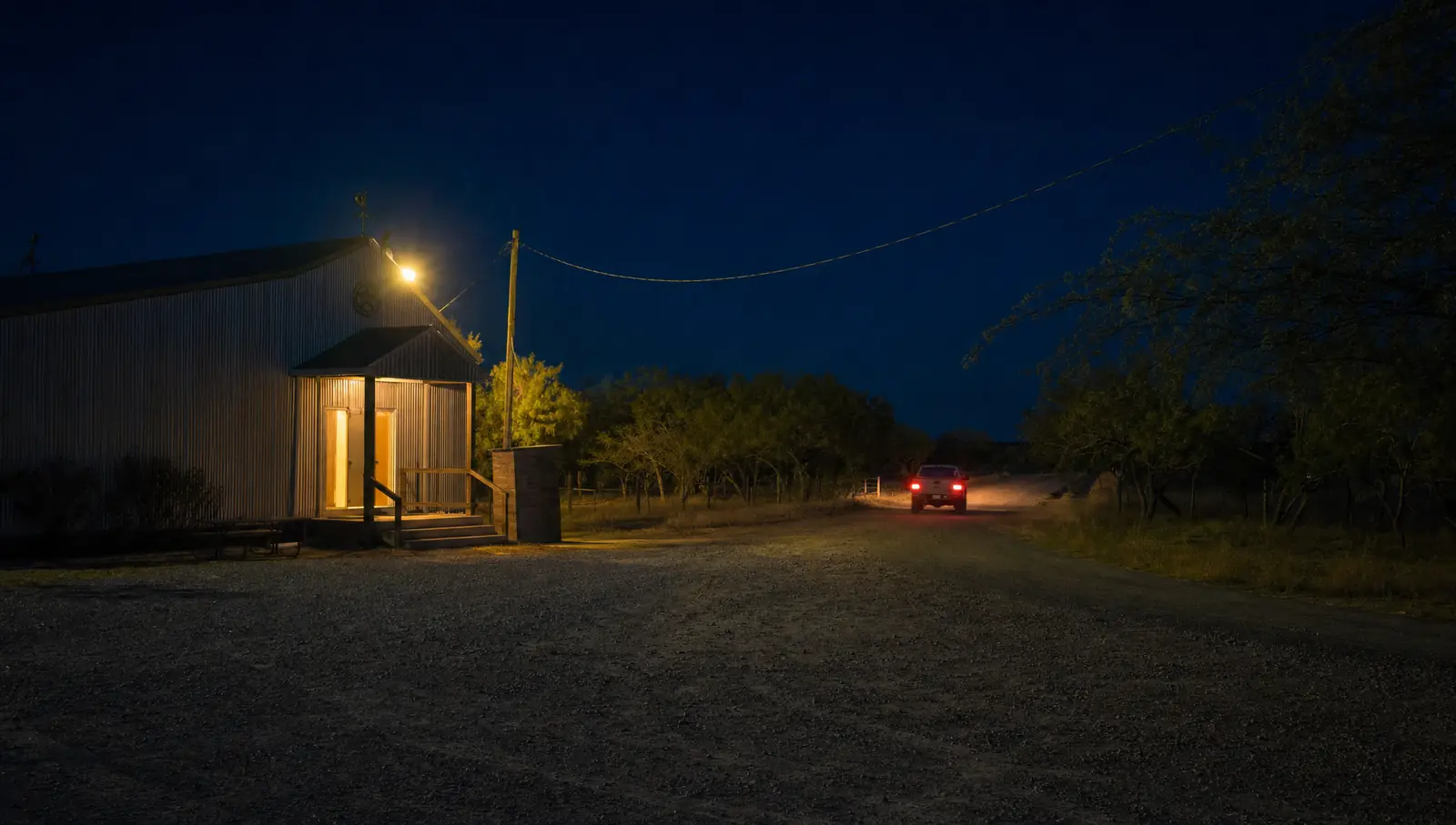 Una camioneta con las luces encendidas en una carretera vacía frente a un salón de baile rural, de noche, con un poste de luz anaranjada